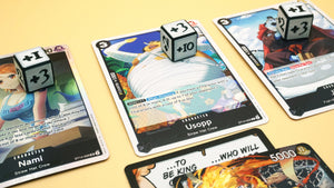 Multiple Positive Dice shown in white with black on top of various black character cards from the One Piece TCG
