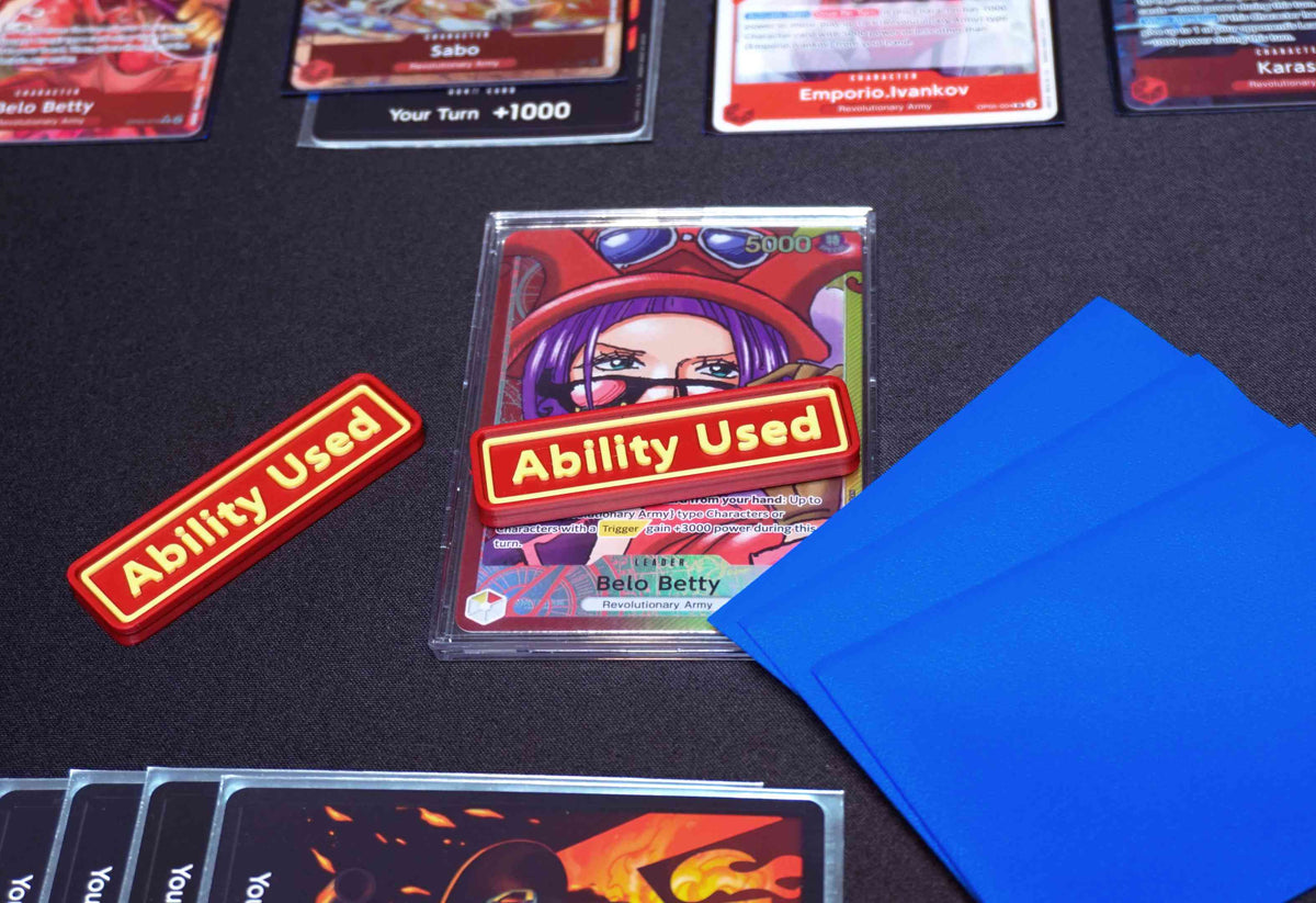 A Red and Yellow Ability User Marker sits on top of the Belo Betty leader alt art card with the hand face down next to it, all on top of a black playmat